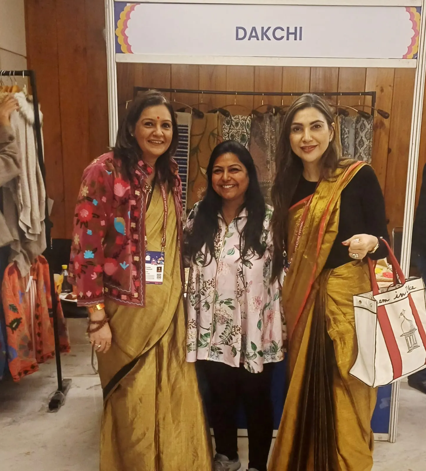 Three women, two in sarees, at the Dakchi stall with textiles on rails behind them
