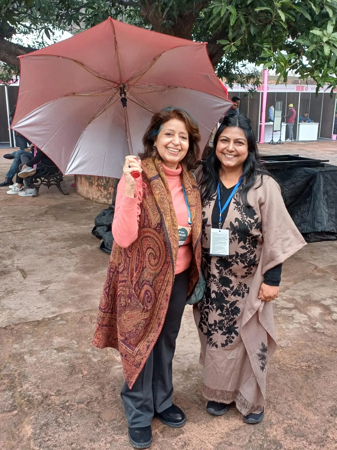 Two women at an outdoor exhibition, one holding a pink umbrella, both wearing event badges