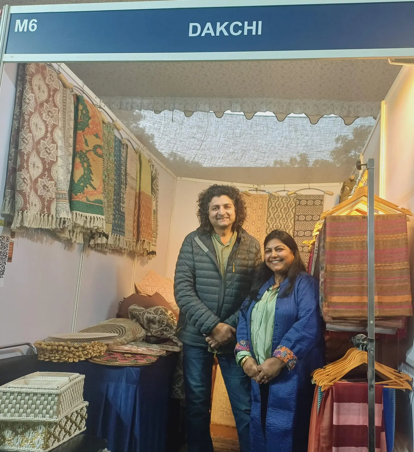 Two people standing inside the Dakchi stall (M6), with rugs on the walls and baskets on the table