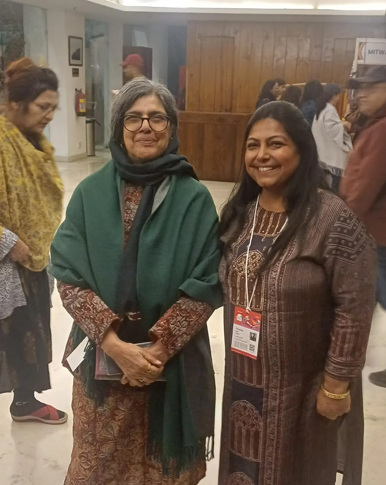 Two women smiling together at an indoor exhibition, both wearing event badges