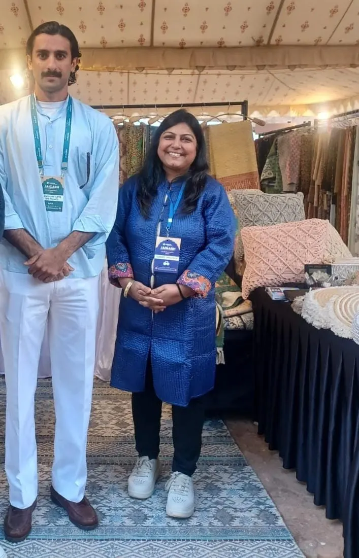 A man in white and a woman in a blue jacket, both wearing event lanyards, at a stall with cushions and rugs