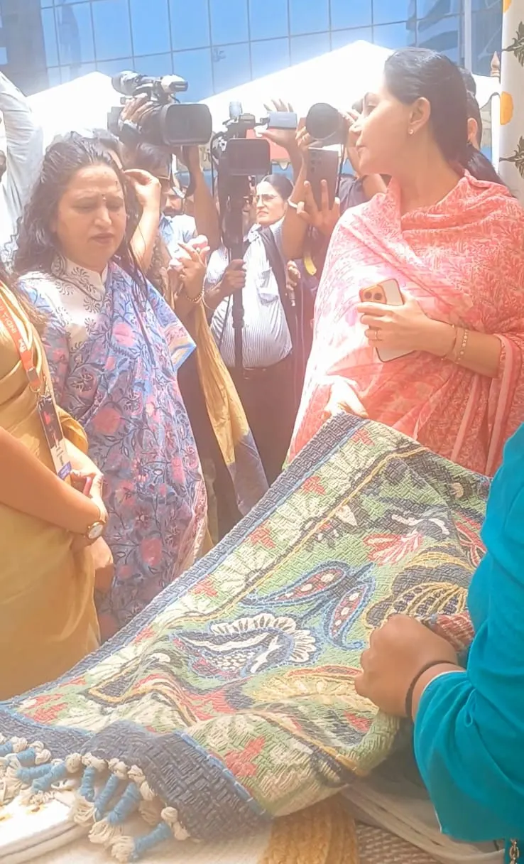 A guest in a coral dupatta looking at a patterned rug held up at the Dakchi stall, with photographers behind