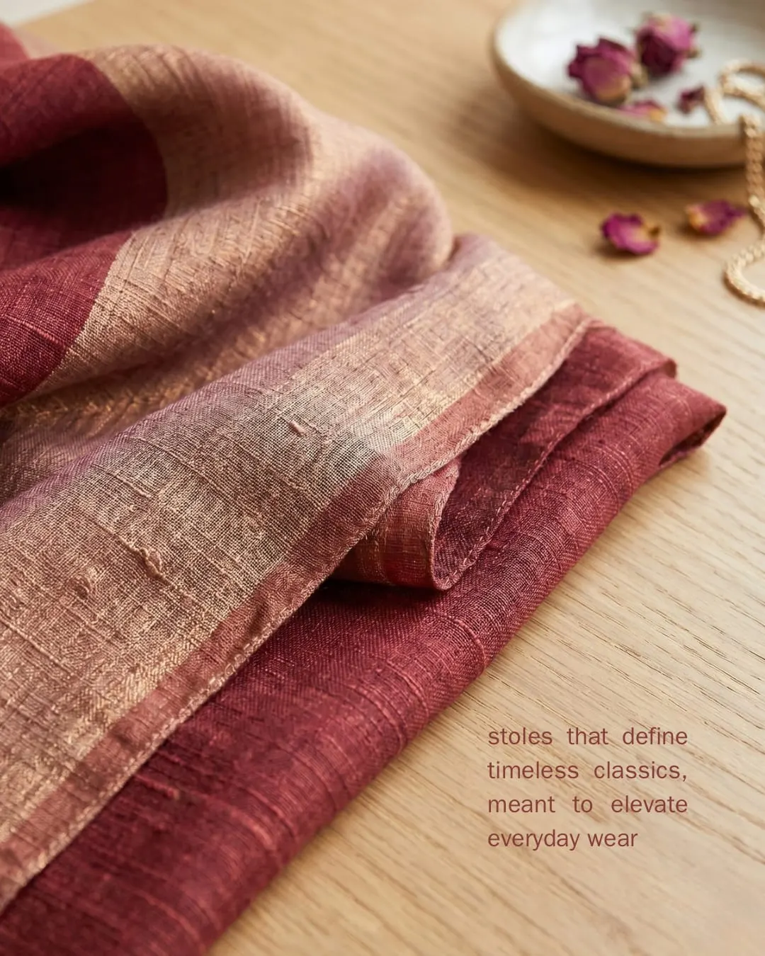 A folded pashmina in deep red and rose on a wooden table with dried rose petals. Text reads Styles that define timeless classics, meant to elevate everyday wear.