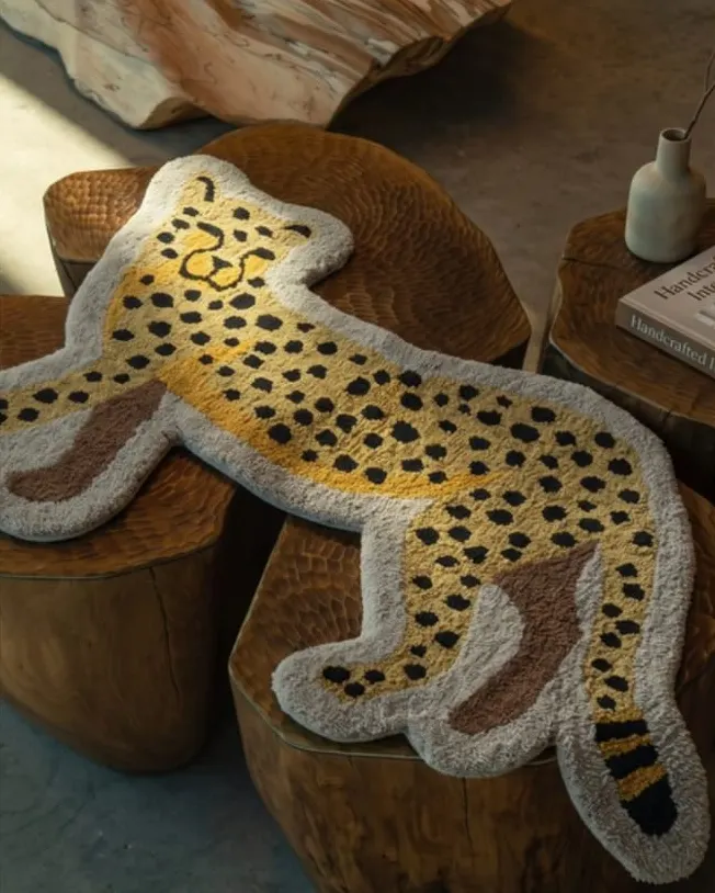 A cheetah-shaped tufted rug in mustard with black spots and a cream border, draped over carved wooden stools