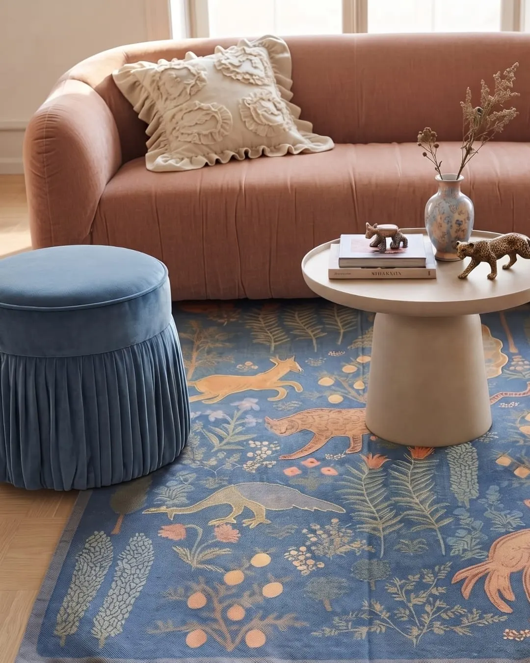 A blue rug with foxes, birds and plants in rust and gold, under a velvet stool and a round side table in a living room