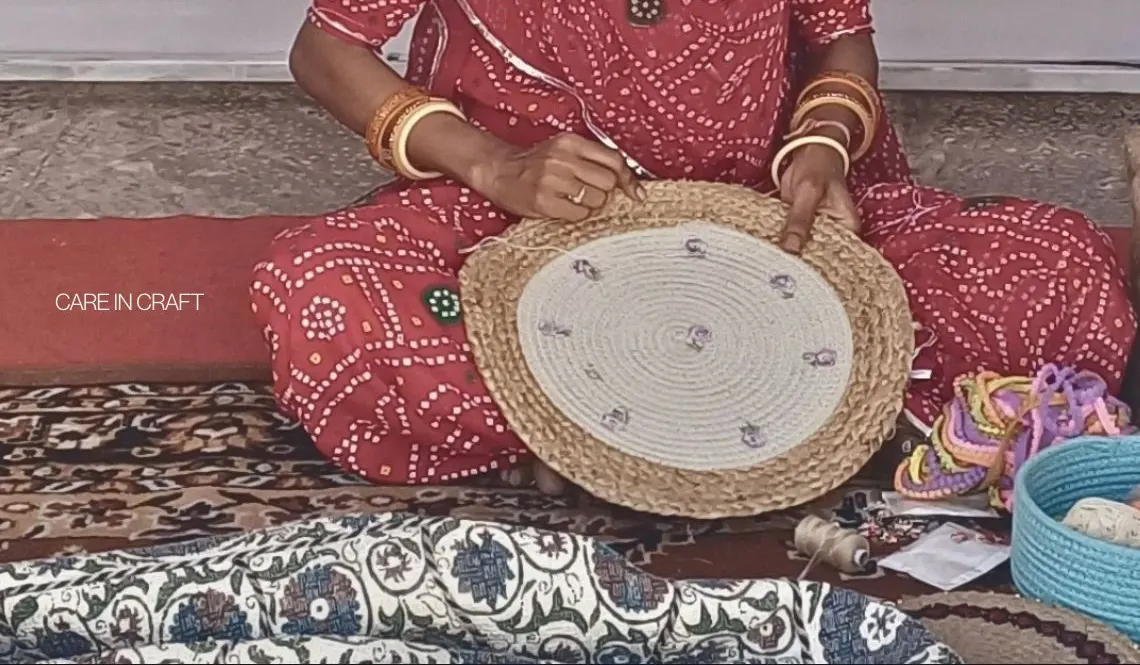 A woman in a red bandhani outfit stitching a round jute and cotton piece by hand, with thread and baskets beside her. Text reads Care in craft.